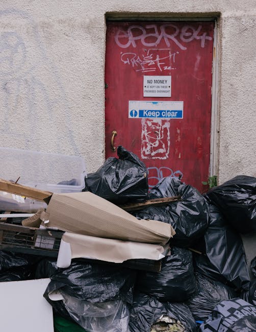 A person wearing a white glove and a wristwatch is holding a large white bag filled with garden debris, including small branches, leaves, and gravel. The bag is situated outdoors on a gravel or dirt s
