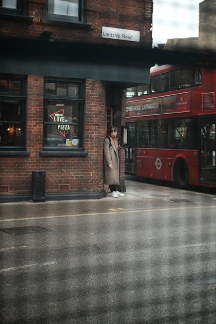 A person wearing a long beige coat, white sneakers, and carrying a bag, stands on the sidewalk outside a brick building, preparing to load or unload items during a home relocation. Behind them, a doub