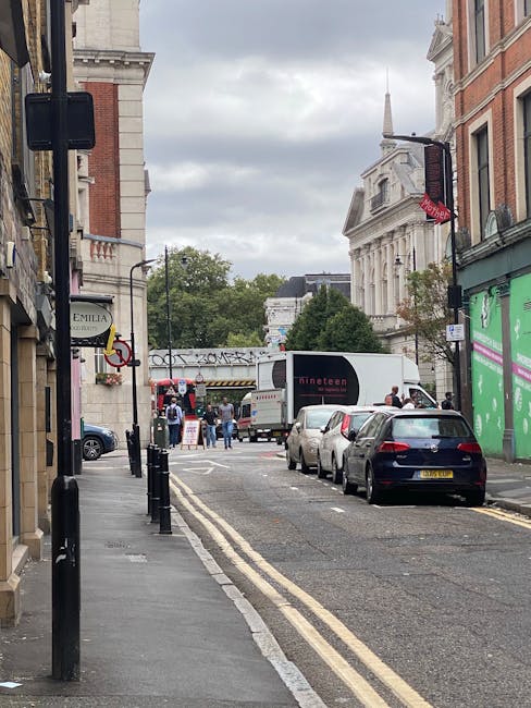 A street scene in Haringey during daytime shows a narrow road lined with parked cars on the right side, including a dark blue vehicle closest to the camera. On the left, there is a sidewalk with black