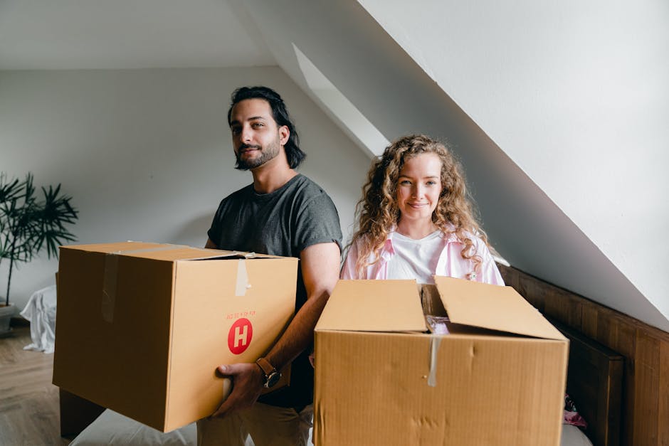 A man with curly hair, wearing a blue t-shirt and dark pants, is sitting on a wooden floor inside a room with large arched windows. Several cardboard boxes, some sealed with red packing tape, are stac