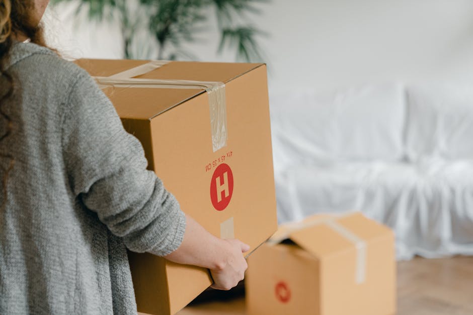 A young couple is sitting on a white sofa in a living room with light-colored wooden flooring and plain white walls during their house move. The woman has curly hair and is wearing a gray cardigan and