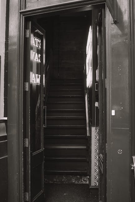 A narrow wooden staircase inside a property, visible through an open doorway, with unfinished, weathered wooden steps and rough wooden framing around the entrance. The surrounding walls are exposed, d